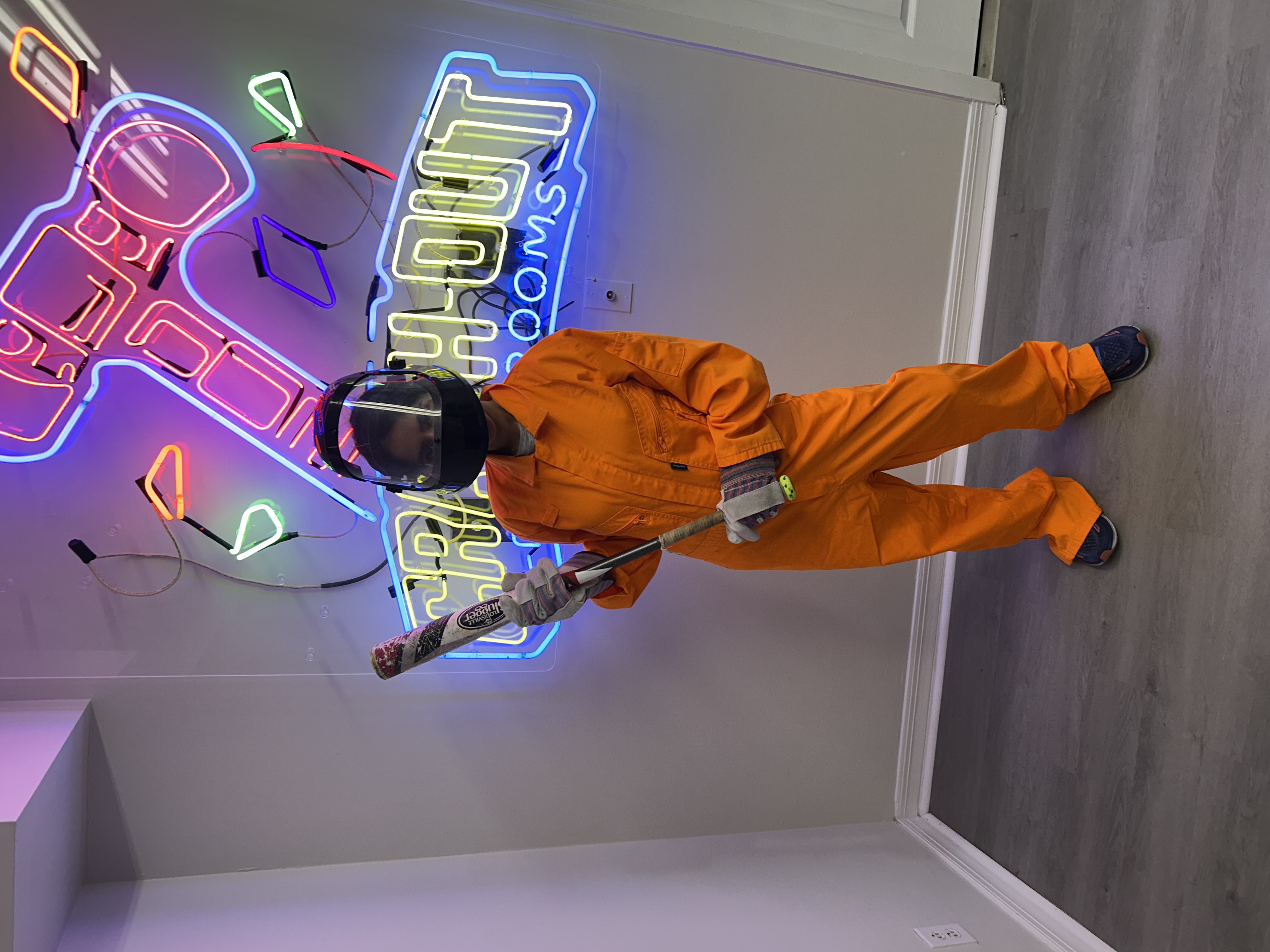 Crash Out customer in full orange jumpsuit and face protection posing with bat in front of Crash Out neon sign