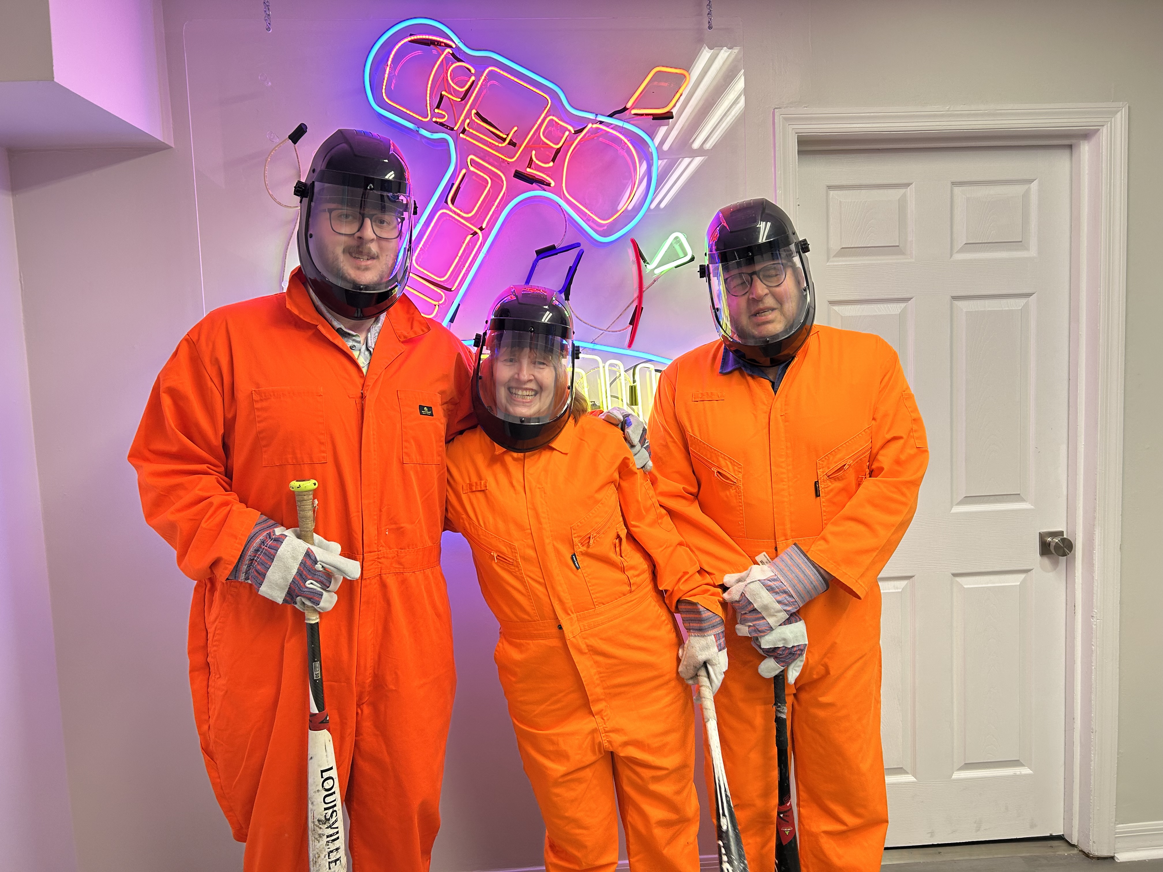 Three Crash Out guests smiling in orange jumpsuits and face shields ready to enter the smash room