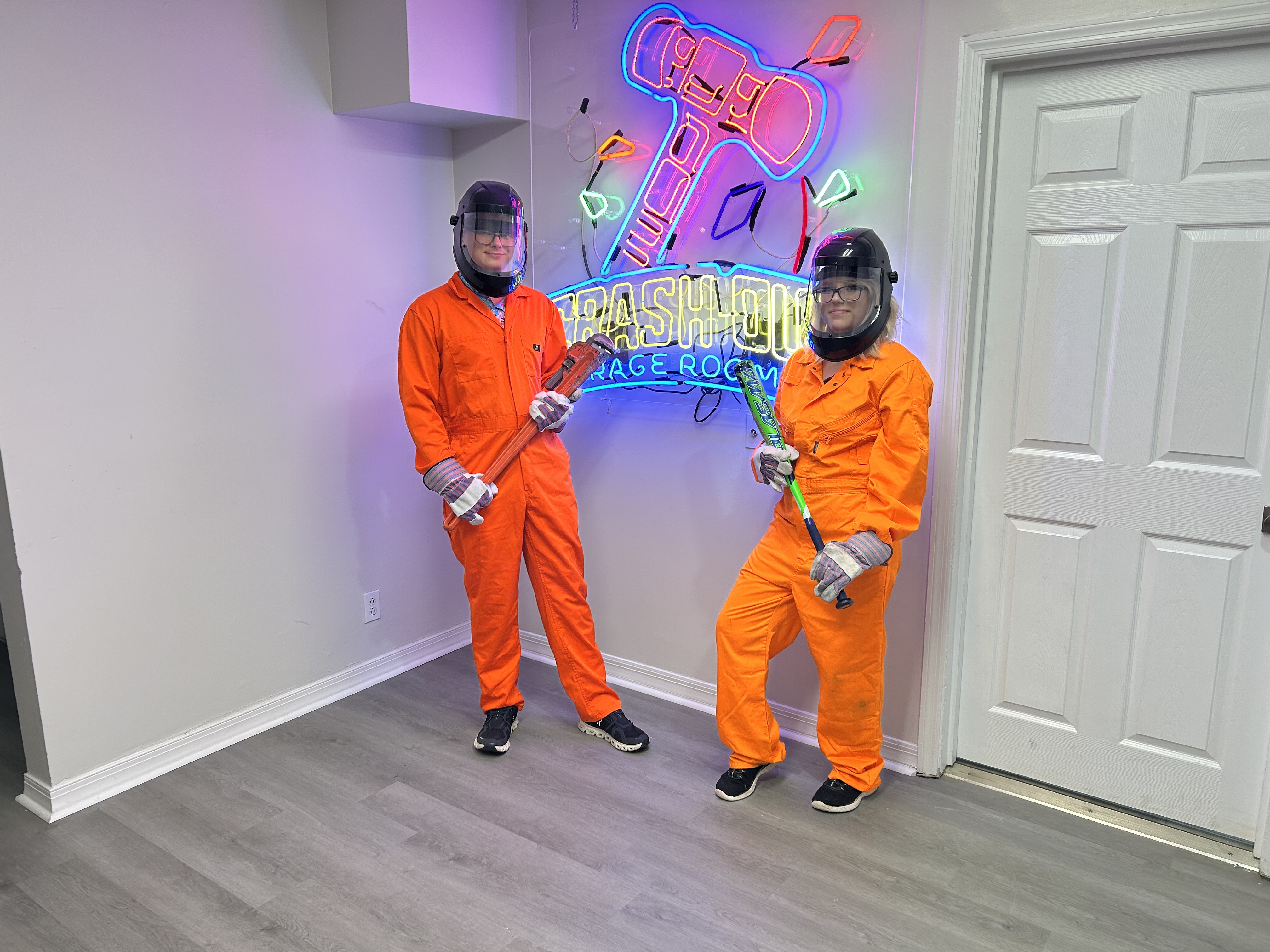 Large group of Crash Out Rage Rooms guests in orange jumpsuits holding bats and smash tools in front of neon sign