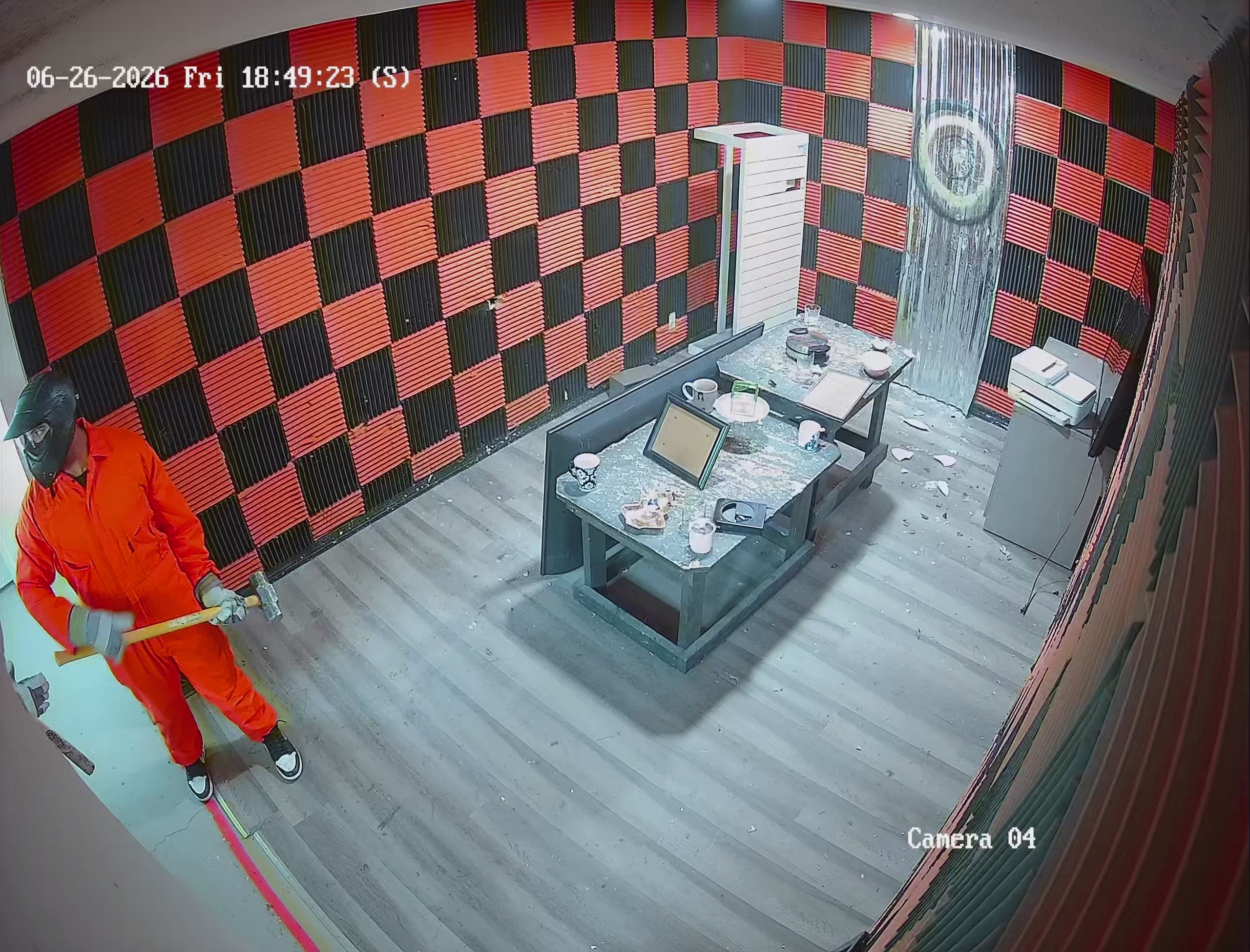 Security camera view of Crash Out Rage Rooms guest in orange jumpsuit swinging sledgehammer inside red and black checkered smash room